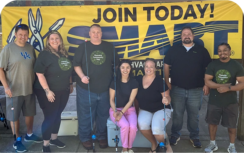 A group of seven people stands in front of a bright yellow banner promoting “Join Today! SMART,” all wearing matching T-shirts for Human Community Day