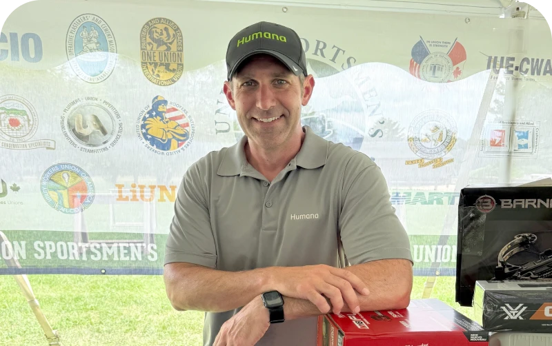 A man in a gray Humana polo and cap stands in front of a tent displaying various sporting event logos and merchandise
