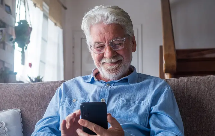 An older person wearing a blue shirt sits comfortably, holding a smartphone and looking at it intently