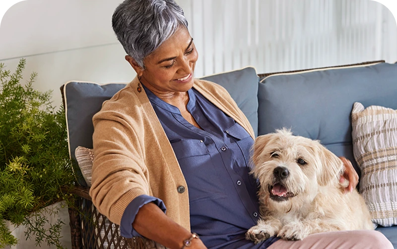 An elderly woman relaxes on a cozy couch, gently petting a fluffy dog
