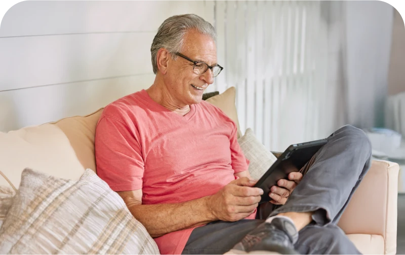 An older man in a t-shirt sits comfortably on a couch, engaged with a tablet, surrounded by soft cushions