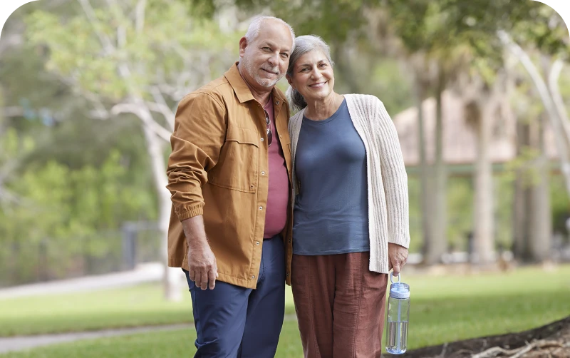 An older couple stands closely together in a park, surrounded by green trees