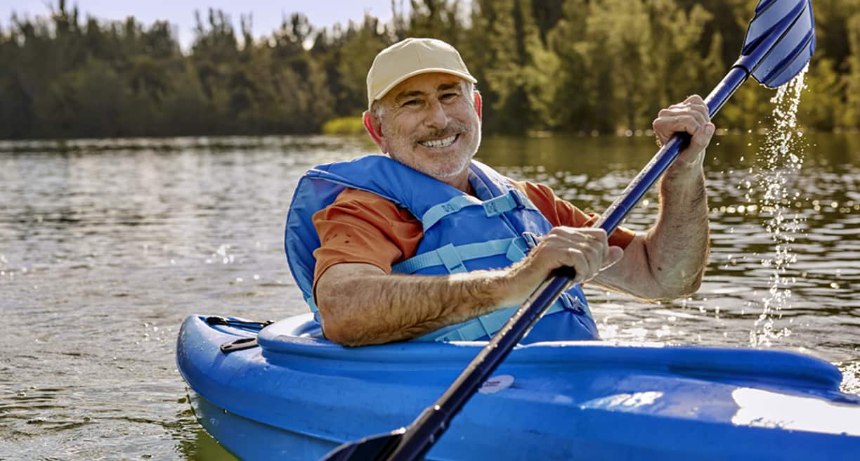 A smiling man paddles a blue kayak on a serene lake, wearing a life vest and cap