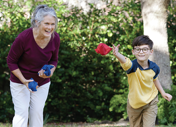 A child in a yellow and blue shirt throws a red beanbag, while an elderly woman holds blue beanbags, both outdoors