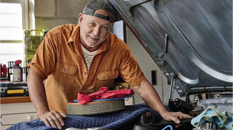 Man in a workshop leans over an open car hood, smiling warmly