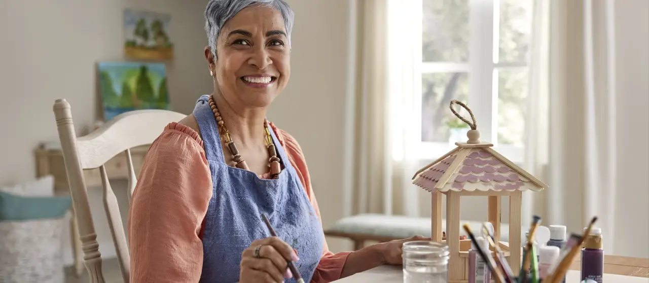 A person with short, silver hair sits at a table, painting on a canvas, surrounded by art supplies