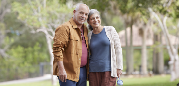An elderly couple smiles together outdoors, surrounded by greenery showcasing an affectionate moment