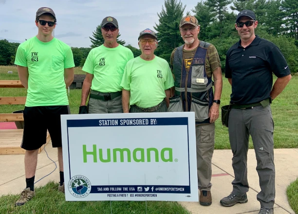 Five men stand together outdoors, wearing matching green shirts, beside a sign displaying “Humana.”