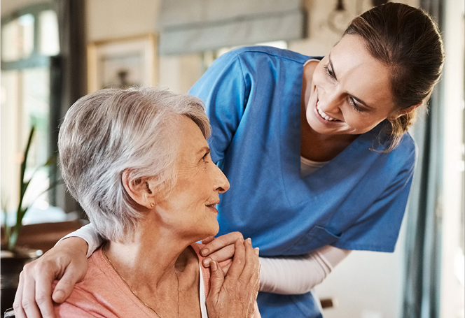 A smiling caregiver in blue scrubs gently rests her hands on an elderly woman's shoulders