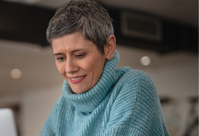 Smiling gray-haired woman in blue sweater working on laptop with pen and coffee cup nearby