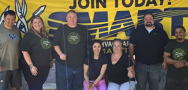 A group of seven people stands in front of a bright yellow banner promoting 'Join Today! SMART,' all wearing matching T-shirts for Human Community Day