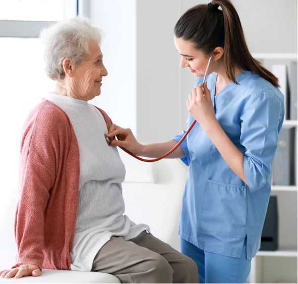 A nurse in blue scrubs uses a stethoscope to check an elderly patient's heartbeat