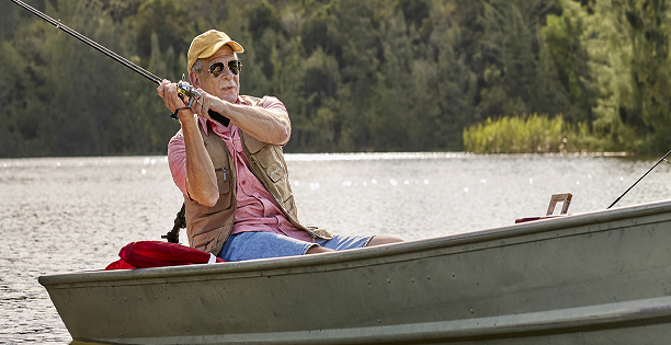 A person in a boat is fishing with a rod on a calm lake, surrounded by lush greenery and peaceful water reflections