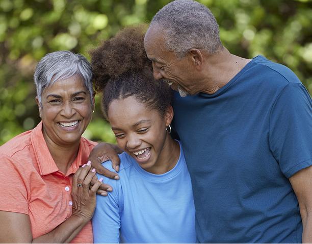 Two older adults and a teenage girl stand close together outdoors, smiling and laughing, with greenery in the background