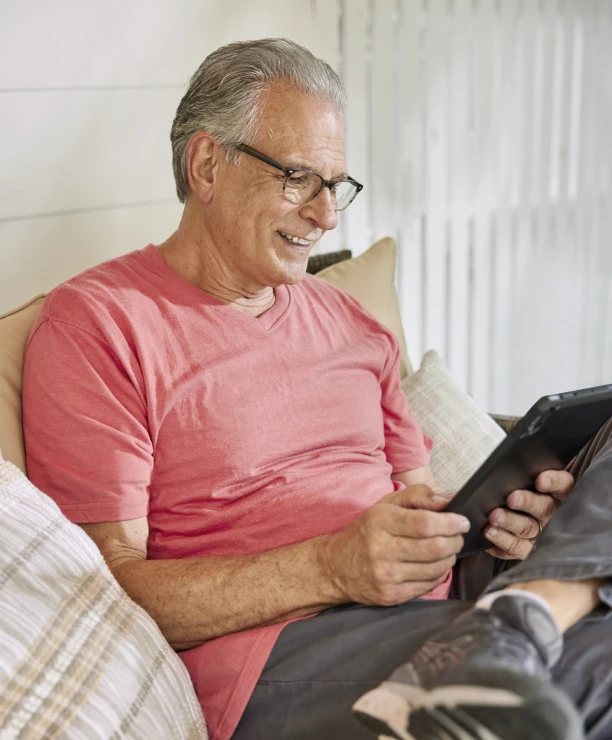 An elderly man in a red shirt uses a tablet outdoors, surrounded by a white picket fence
