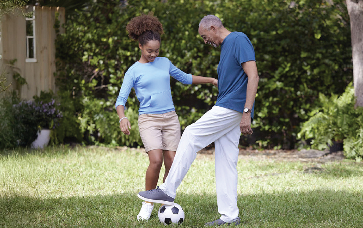 An adult and a child play soccer together on grass, smiling and enjoying their time in a sunny backyard