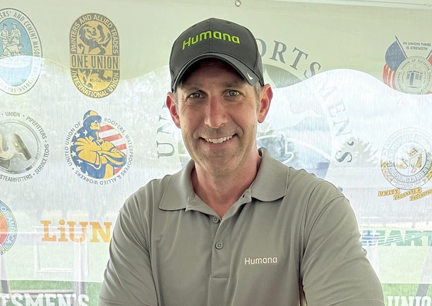A person in a grey shirt and hat stands beside a red box labeled “FUEL” at an outdoor event with various logos in the background