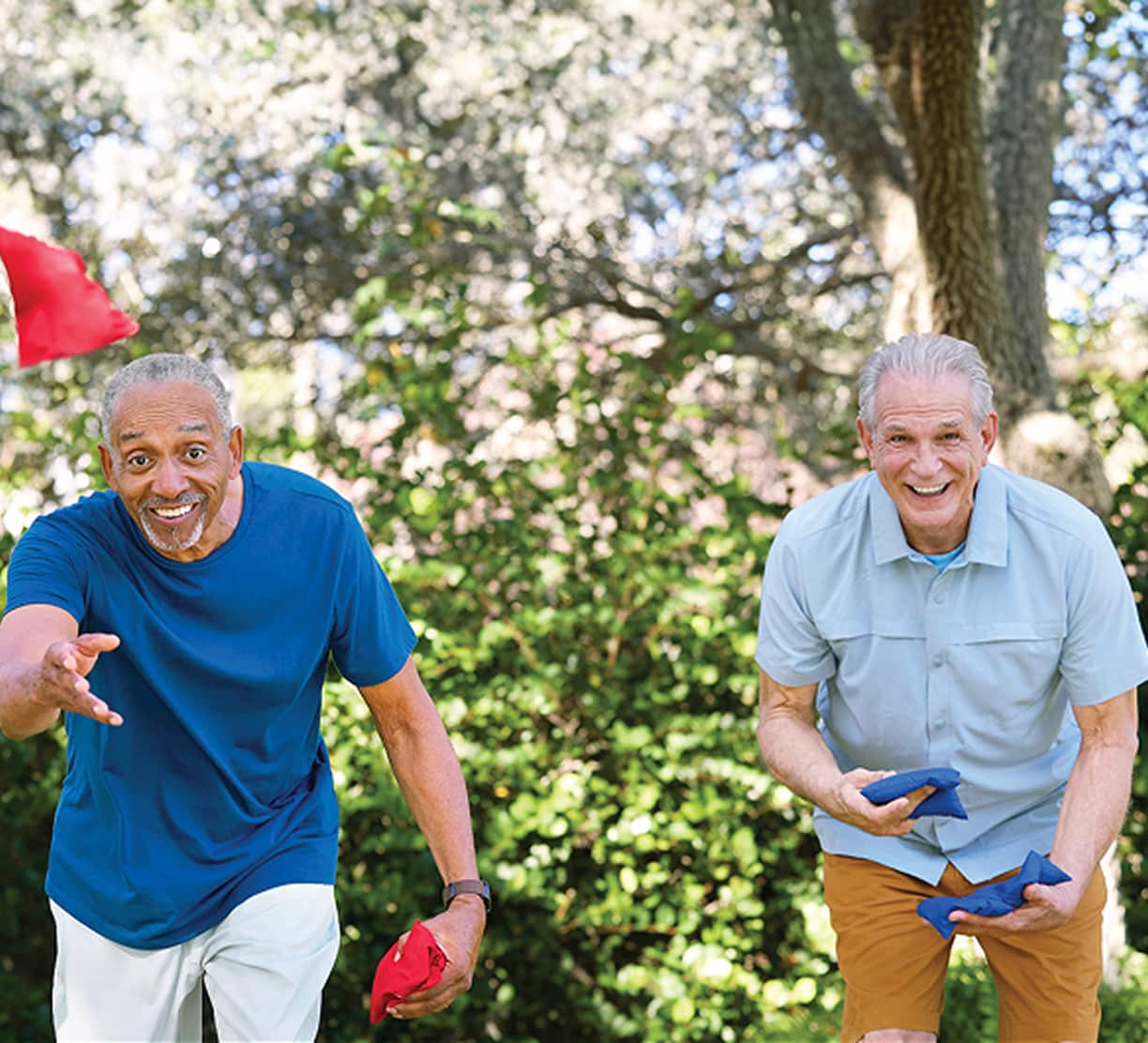 Two smiling older men play cornhole in a sunny park, surrounded by trees. One throws a red beanbag, the other holds blue ones, enjoying the game