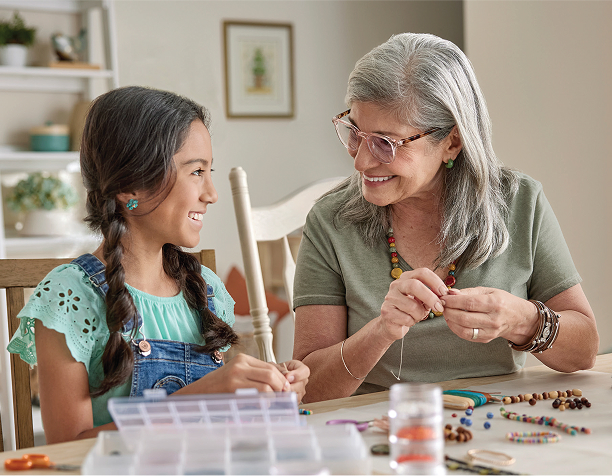 Senior women and a young girl bead crafting at a table, smiling warmly. Surrounded by beads, tools, and containers, they create a joyful atmosphere