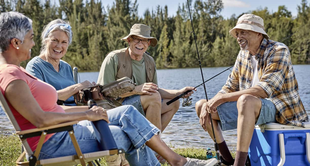 A group of adults enjoys a sunny day by a lake, engaged in conversation while fishing, surrounded by trees and water