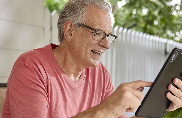 An elderly man in a red shirt uses a tablet outdoors, surrounded by a white picket fence