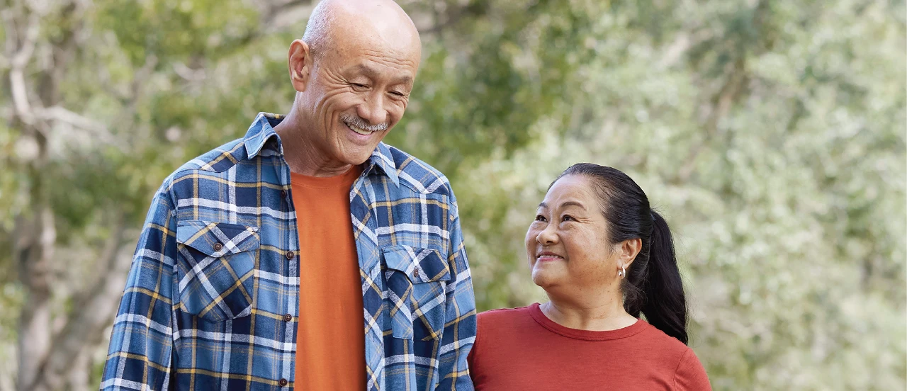 A man and a woman standing together outdoors, smiling, dressed casually in a forested area background