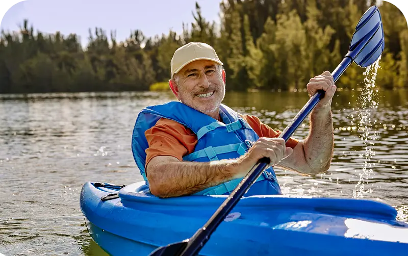 A person paddles a blue kayak on a calm lake, surrounded by lush greenery under bright sunlight. Water splashes from the paddle