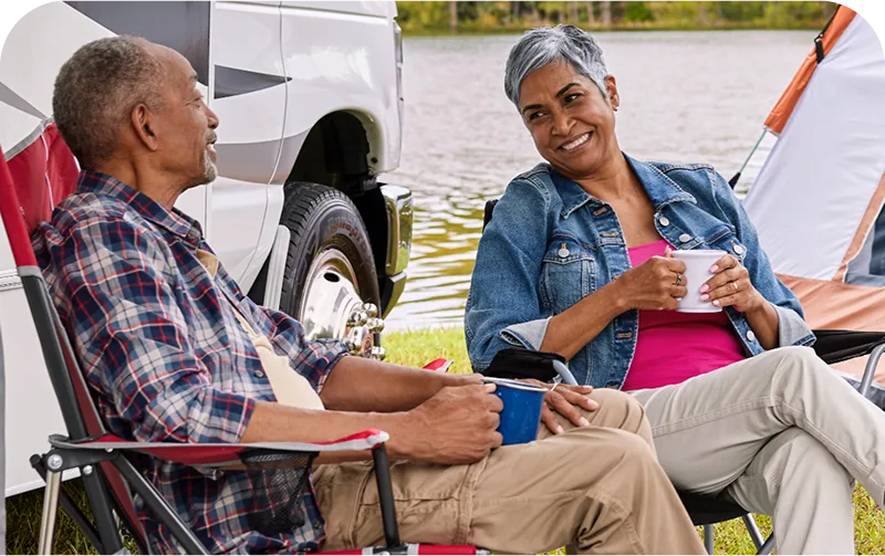A couple relaxing by a lakeside in camping chairs, enjoying drinks, with a camper van and tent in the background