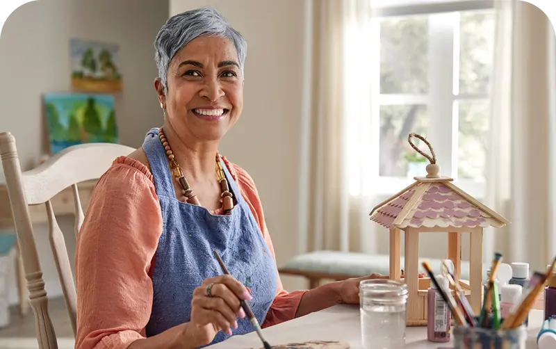 An artist in an apron paints at a sunlit table, surrounded by art supplies and a decorative lantern. Colorful paintings are displayed in the background.